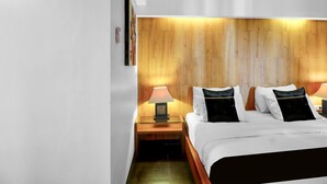 Habitación de hotel con cabecera de madera, cama con ropa blanca y almohadas negras, mesita de noche con lámpara y una obra de arte en la pared.