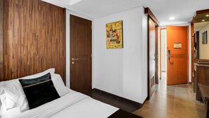 A hotel room with a wooden headboard, a bed with white linens, a door, and a hallway leading to another room. - Palette Hotel Seminyak (Seminyak)