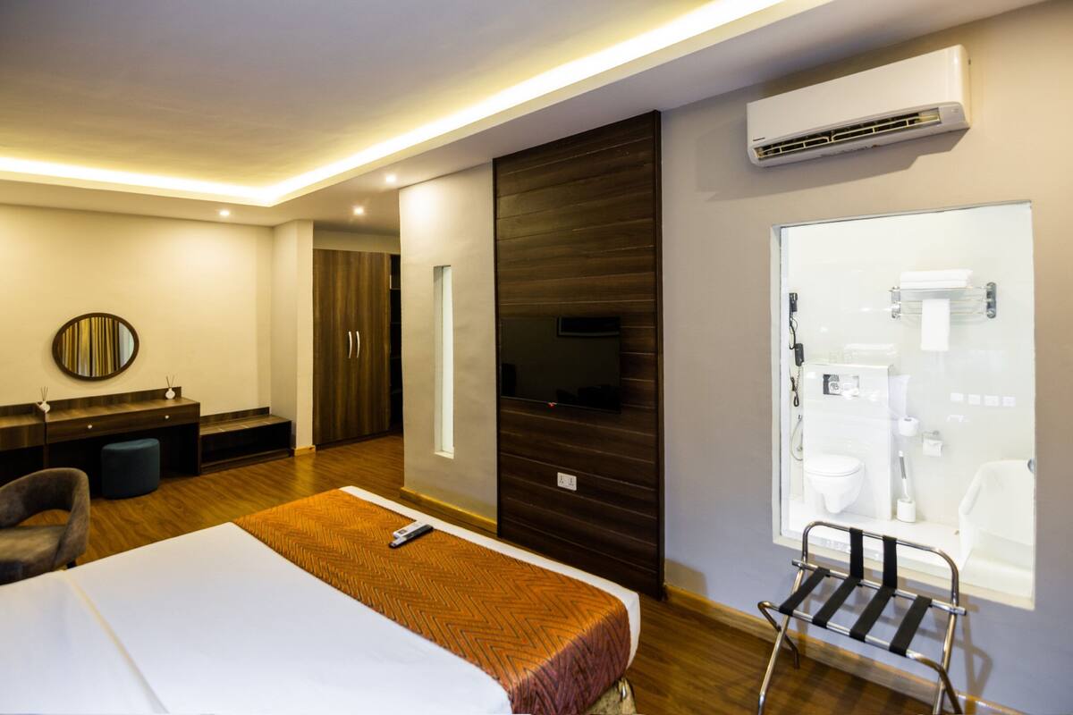 A hotel room with a bed, a desk, a chair, a mirror, a wardrobe, and a bathroom visible through a glass door.