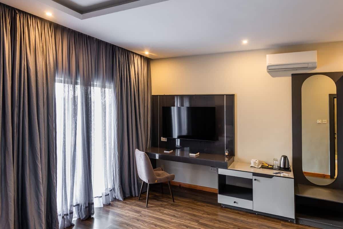 A modern hotel room with a large flat-screen TV, a desk with a chair, a mirror, and a large window with curtains.