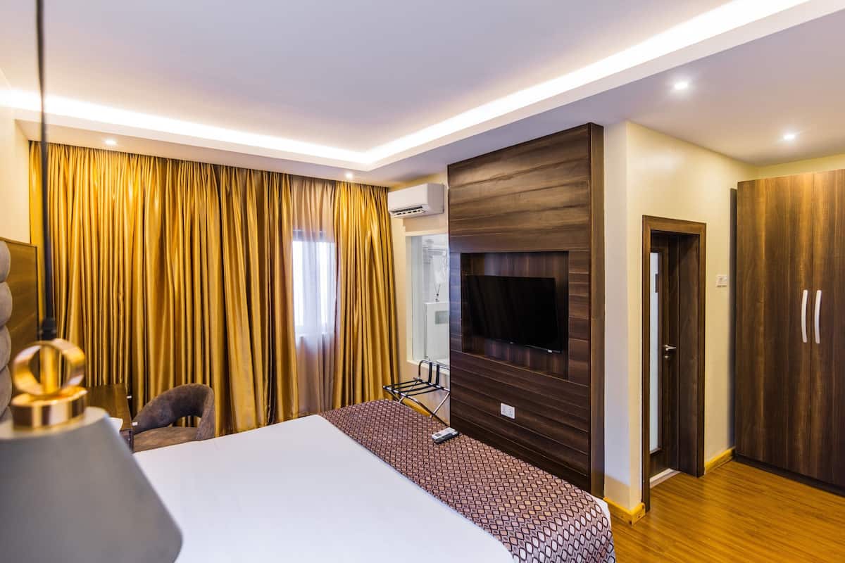 A modern hotel room with a large bed, a flat-screen TV mounted on the wall, a wardrobe, and a seating area with a chair and a small table.