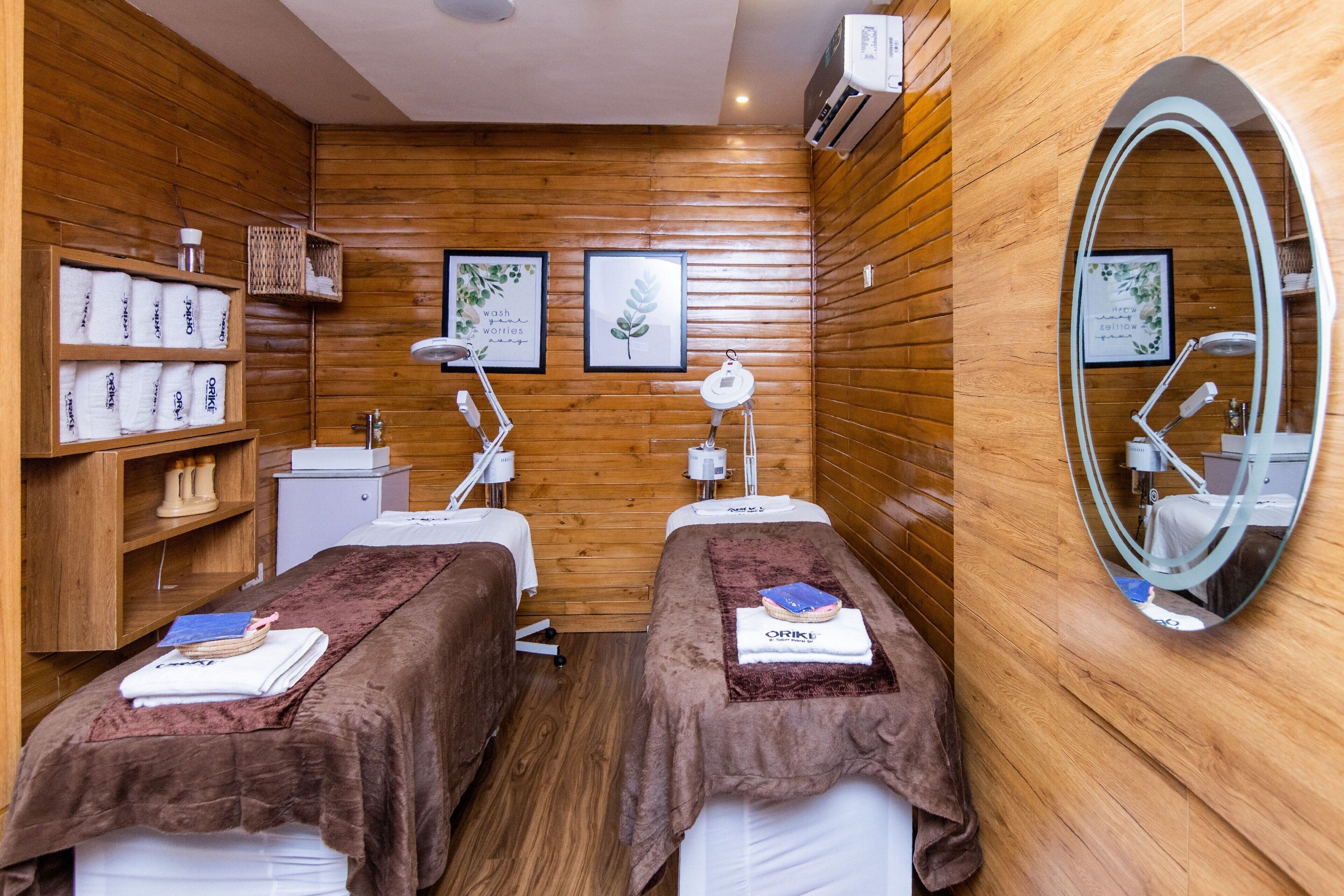 A spa room with wooden walls, two massage tables, a mirror, and shelves with towels and toiletries.