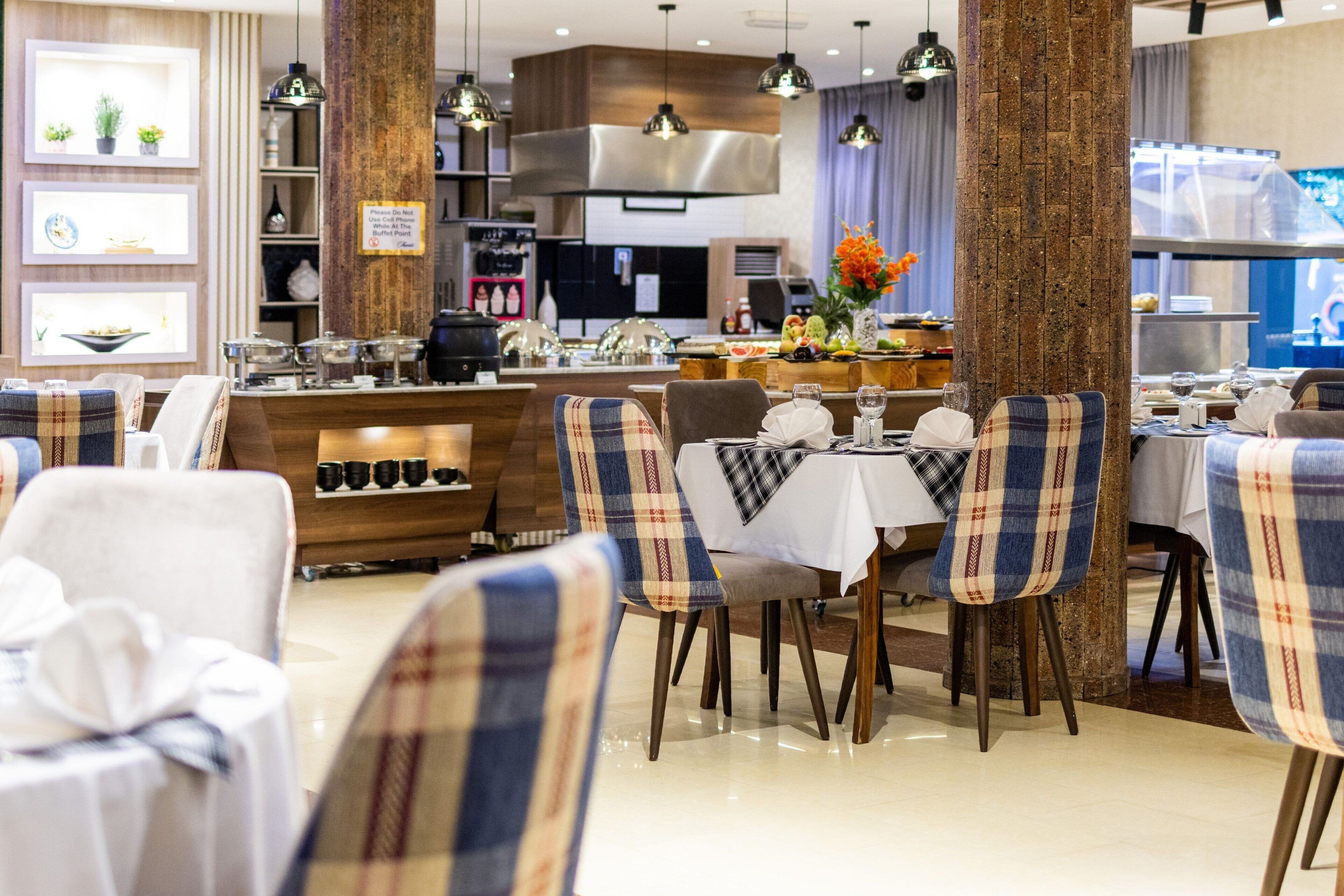 A restaurant with a modern interior, featuring a central dining area with plaid chairs and a white tablecloth. There are shelves with various items and a kitchen area in the background.