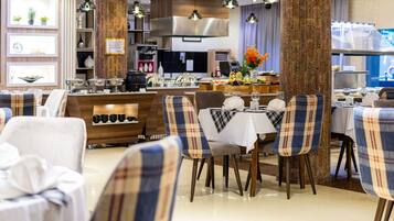 A restaurant with a modern interior, featuring a central dining area with plaid chairs and a white tablecloth. There are shelves with various items and a kitchen area in the background.