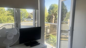 Habitación con televisor sobre un mueble, ventilador blanco y puerta corrediza de vidrio que da a un área exterior con vistas a árboles y un edificio.