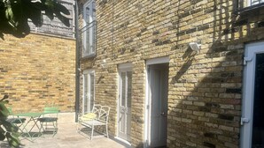 Un bâtiment en briques avec un patio, une table et des chaises vertes, et une porte blanche.