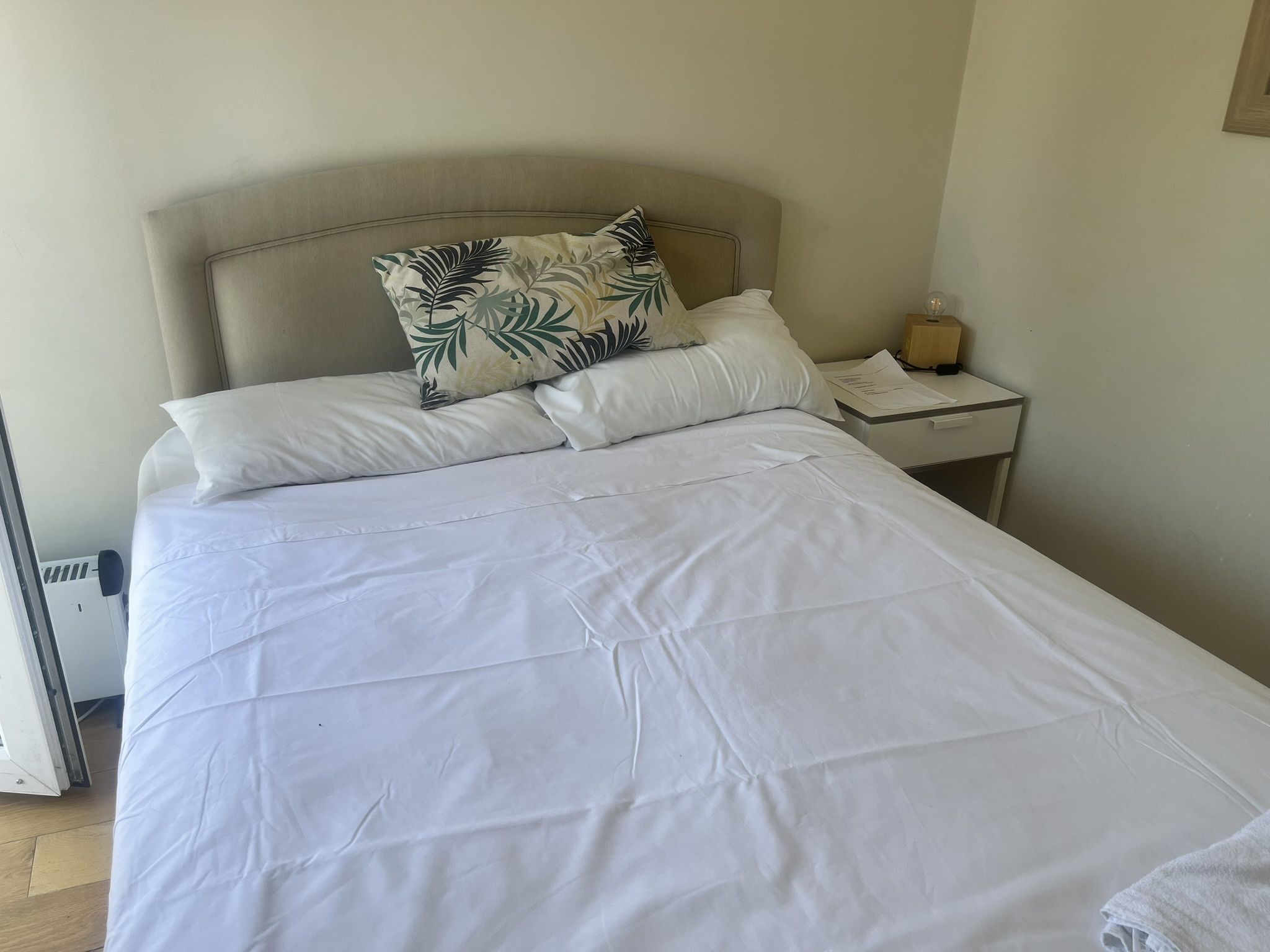 A bed with white bedding and a leaf-patterned pillow, a bedside table with a lamp, and a power outlet on the wall.