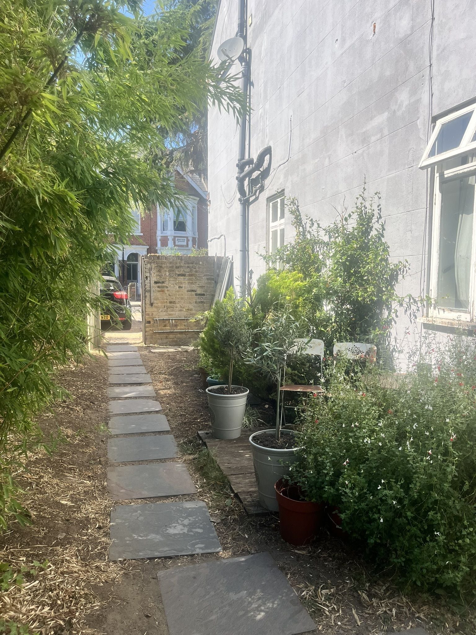 A narrow pathway leading to a building with a brick wall and a window. There are potted plants and a metal structure on the right side.