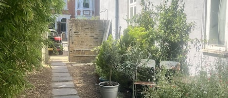 A narrow pathway leading to a building with a brick wall and a window. There are potted plants and a metal structure on the right side.