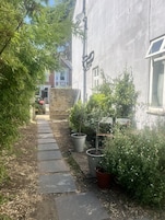 A narrow pathway leading to a building with a brick wall and a window. There are potted plants and a metal structure on the right side.