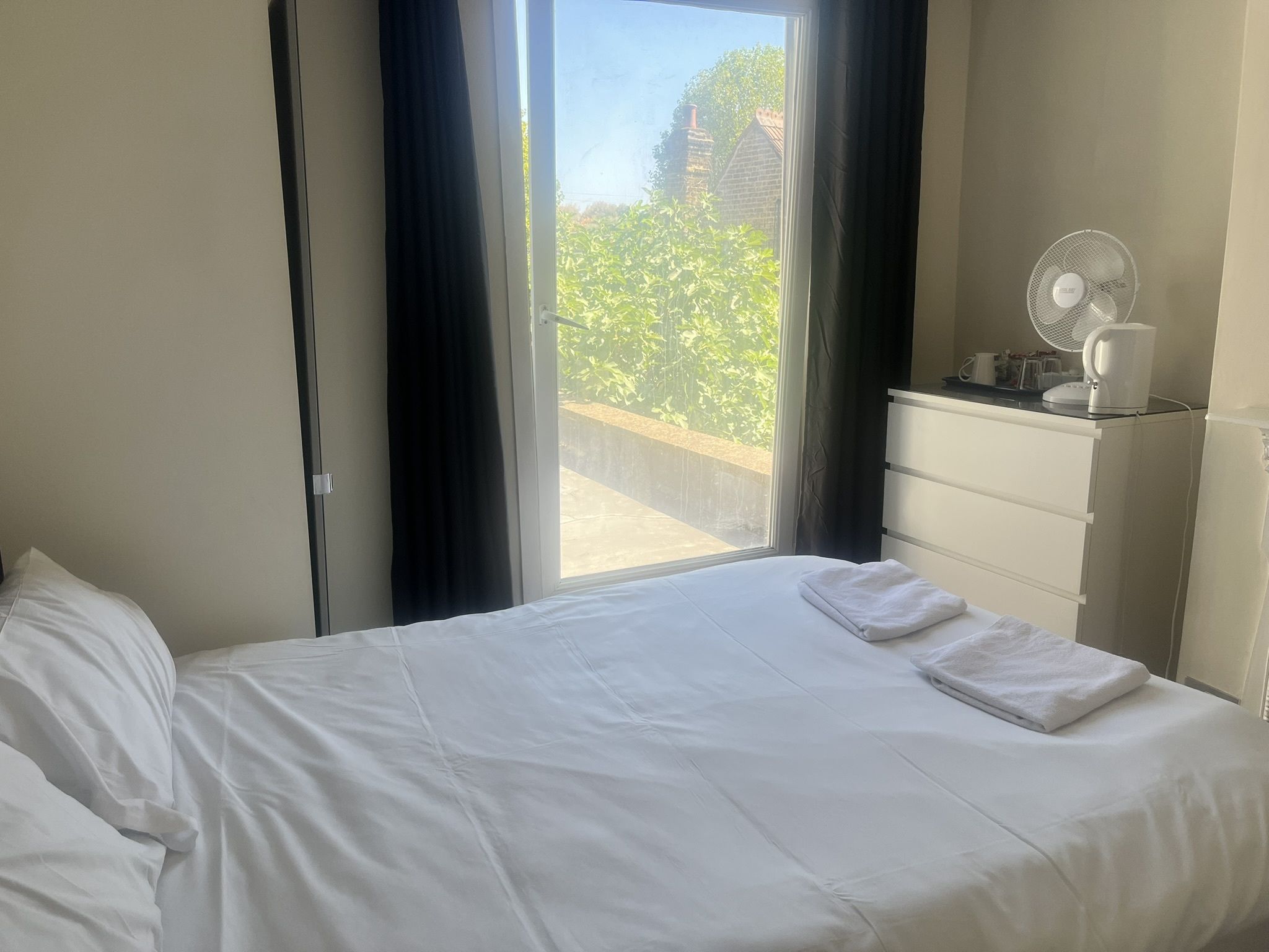 A hotel room with a bed, a desk with a fan, and a view of greenery outside.