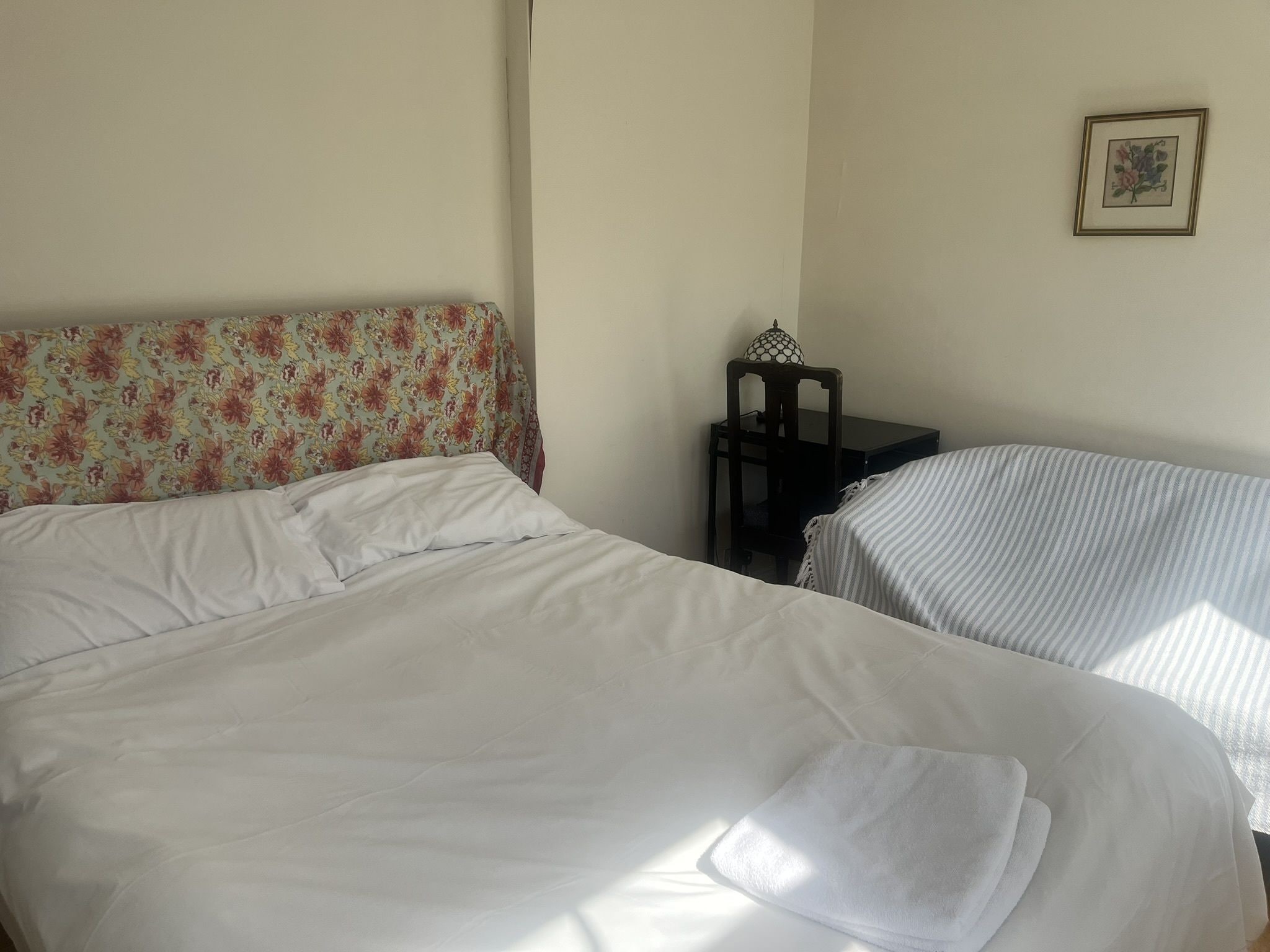 Una habitación con dos camas, una con cabecera de flores y otra con cubrecolcha a rayas. Hay una mesita con una lámpara y una silla.