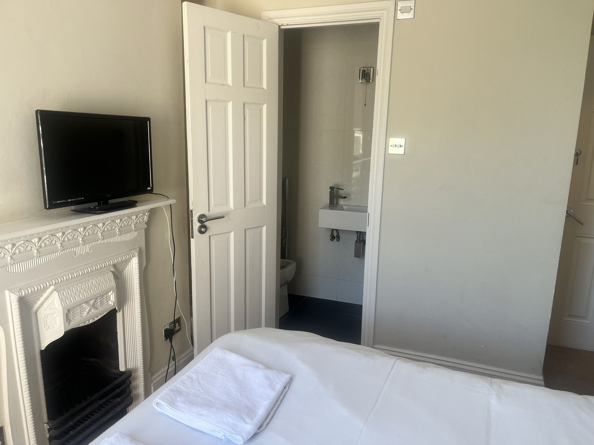 A hotel room with a television on a mantelpiece, a white door leading to a bathroom, and a neatly made bed with a white towel.