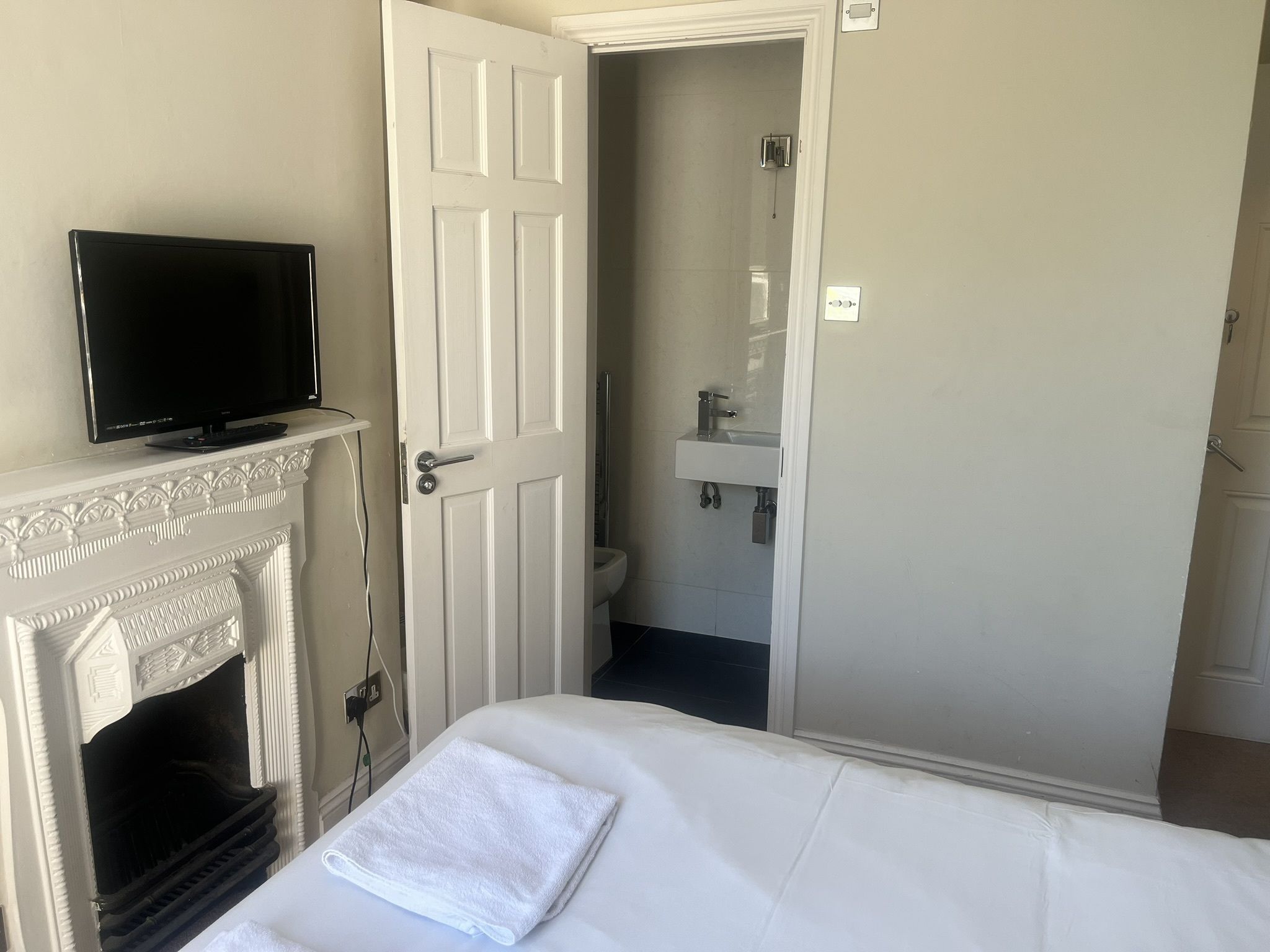 A hotel room with a television on a mantelpiece, a white bed with a folded towel, a door leading to a bathroom, and a white fireplace.