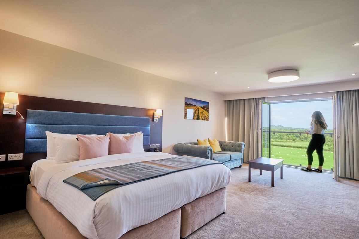 Habitación de hotel con una cama grande, un sofá, una mesita y un balcón con vista a un campo.