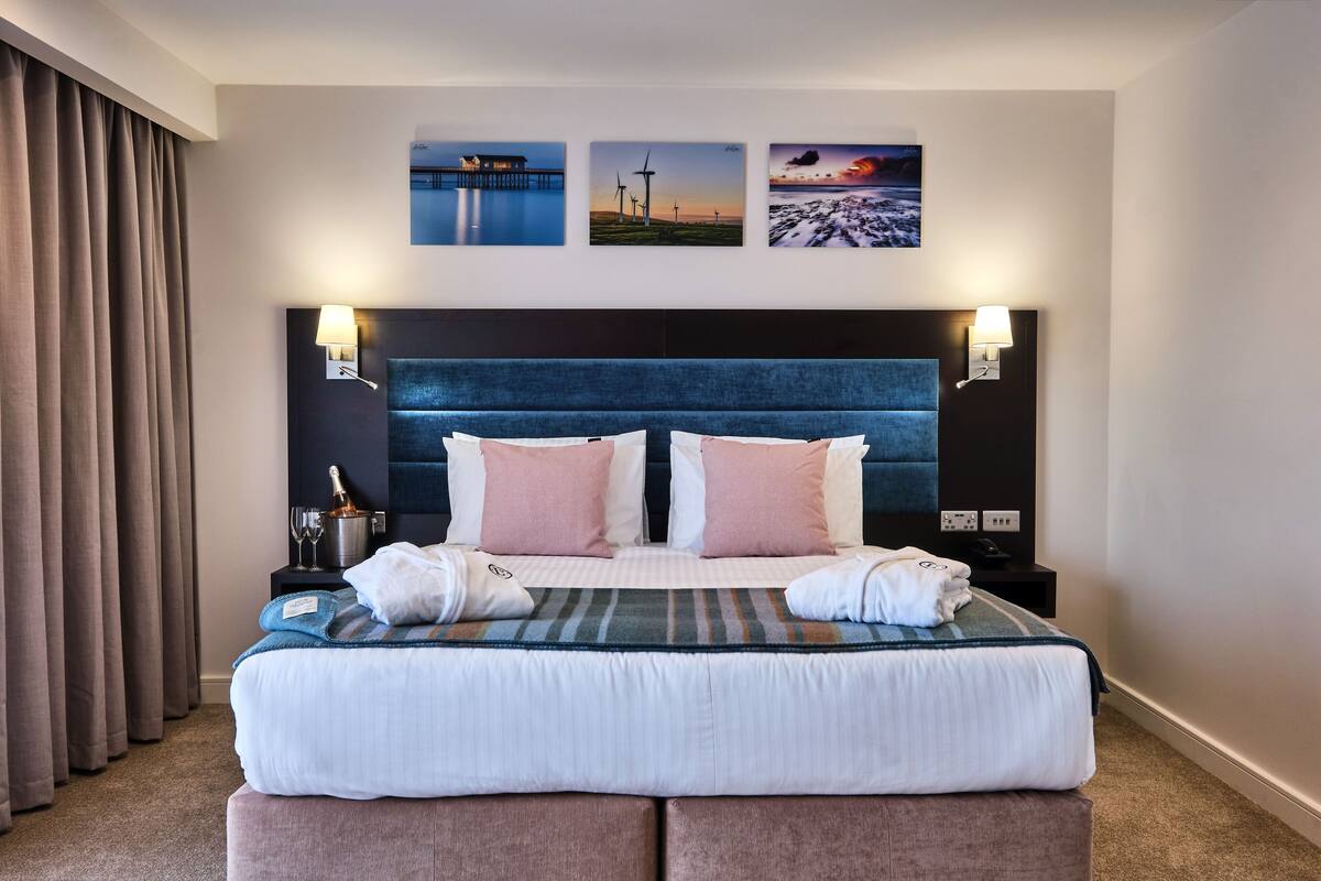 Una cama bien hecha con cabecera azul, dos mesitas de noche con lámparas y tres cuadros en la pared.