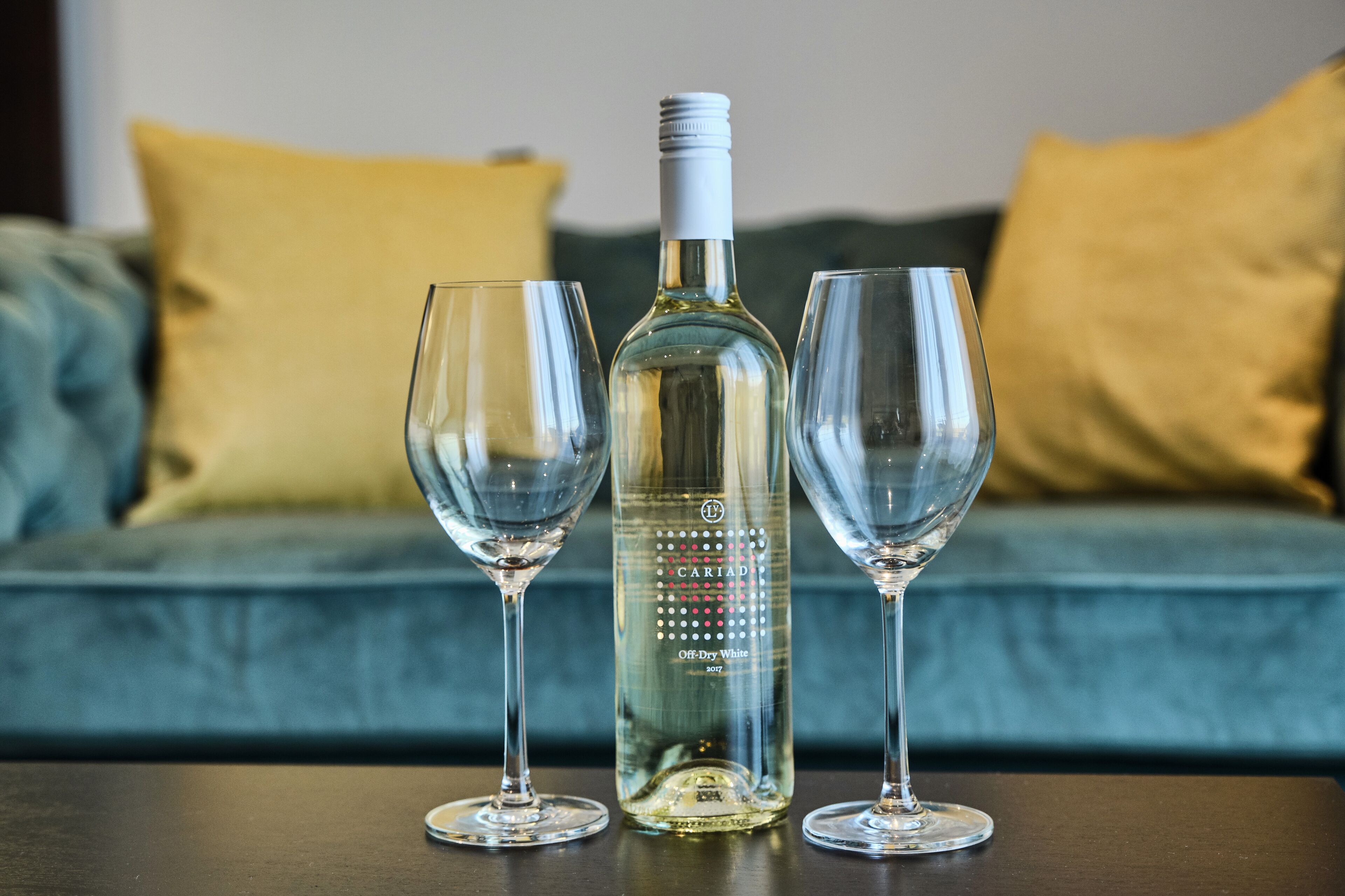 Una botella de vino y dos copas sobre una mesa frente a un sofá con cojines.
