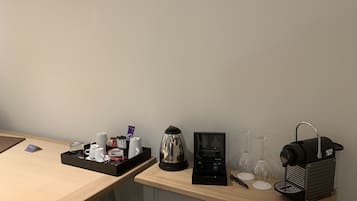 Habitación de hotel con escritorio de madera, cafetera, tetera, copa de vino y una pequeña cesta negra.