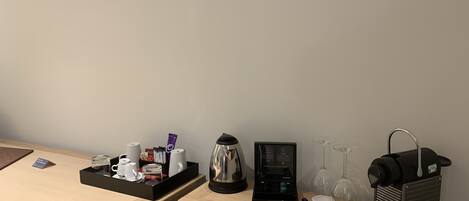 A hotel room with a wooden desk, a coffee maker, a tea kettle, a wine glass, and a small black basket.