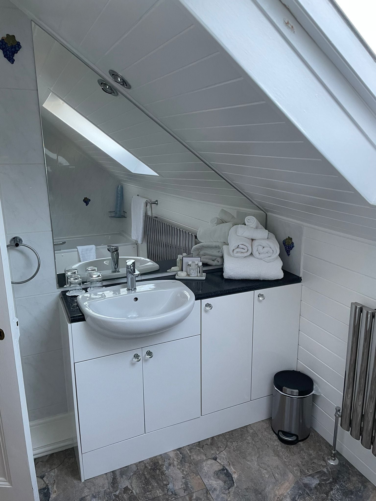 Een moderne badkamer met een wit wastafelmeubel, een wastafel en een spiegel. Er is een handdoekhouder met handdoeken, een prullenbak en een radiator.