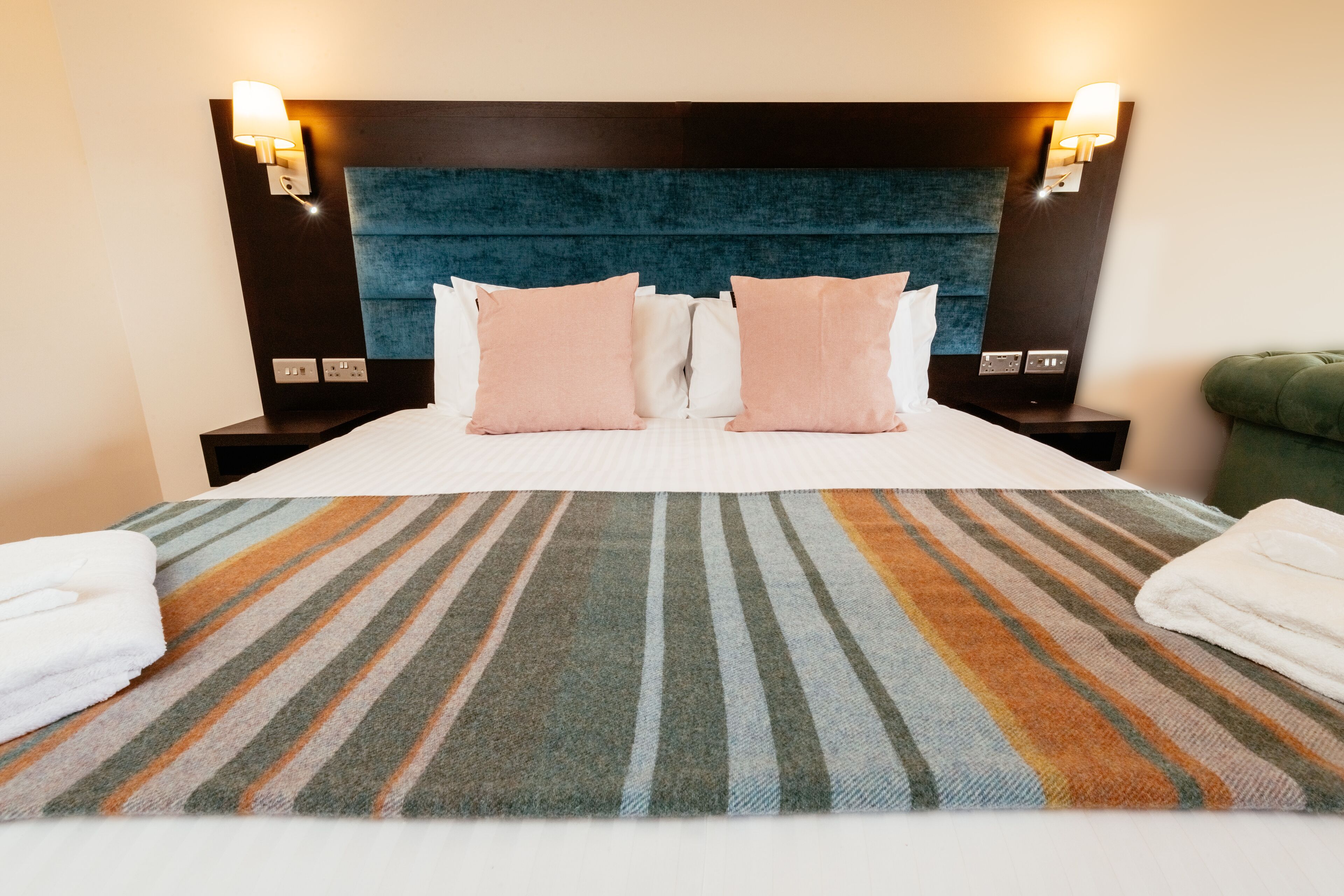 Una cama bien hecha con ropa de cama a rayas, dos mesitas de noche con lámparas y un sillón tapizado en verde.