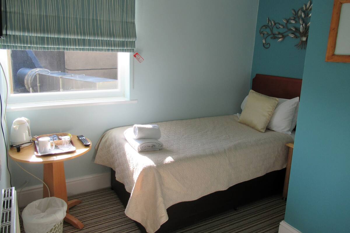 A single bed with a white bedspread, a bedside table with a kettle, a remote control, and a small basket.