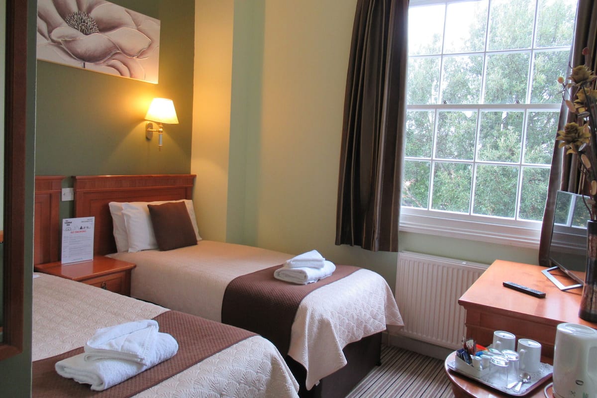 A hotel room with two beds, a nightstand, a desk with a coffee maker, and a window with curtains.