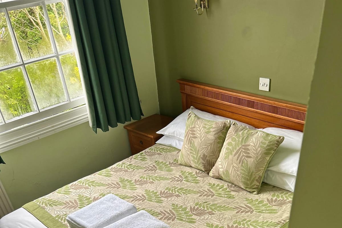 A neatly made bed with a green leaf-patterned bedspread, two white towels, and a wooden headboard.