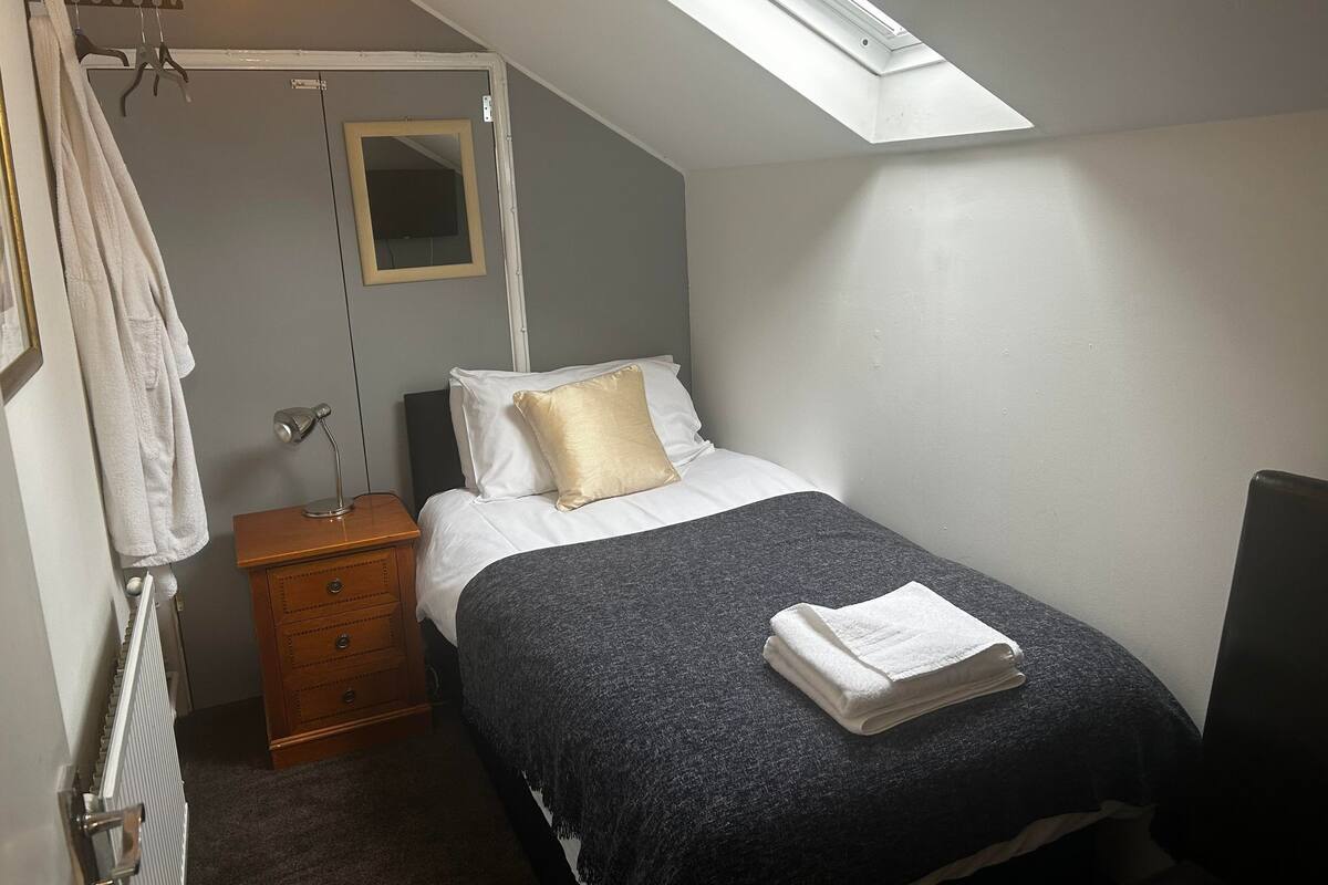 A bedroom with a skylight, a bed with a dark blanket, a bedside table with a lamp, a wooden dresser, and a hanging towel rack.