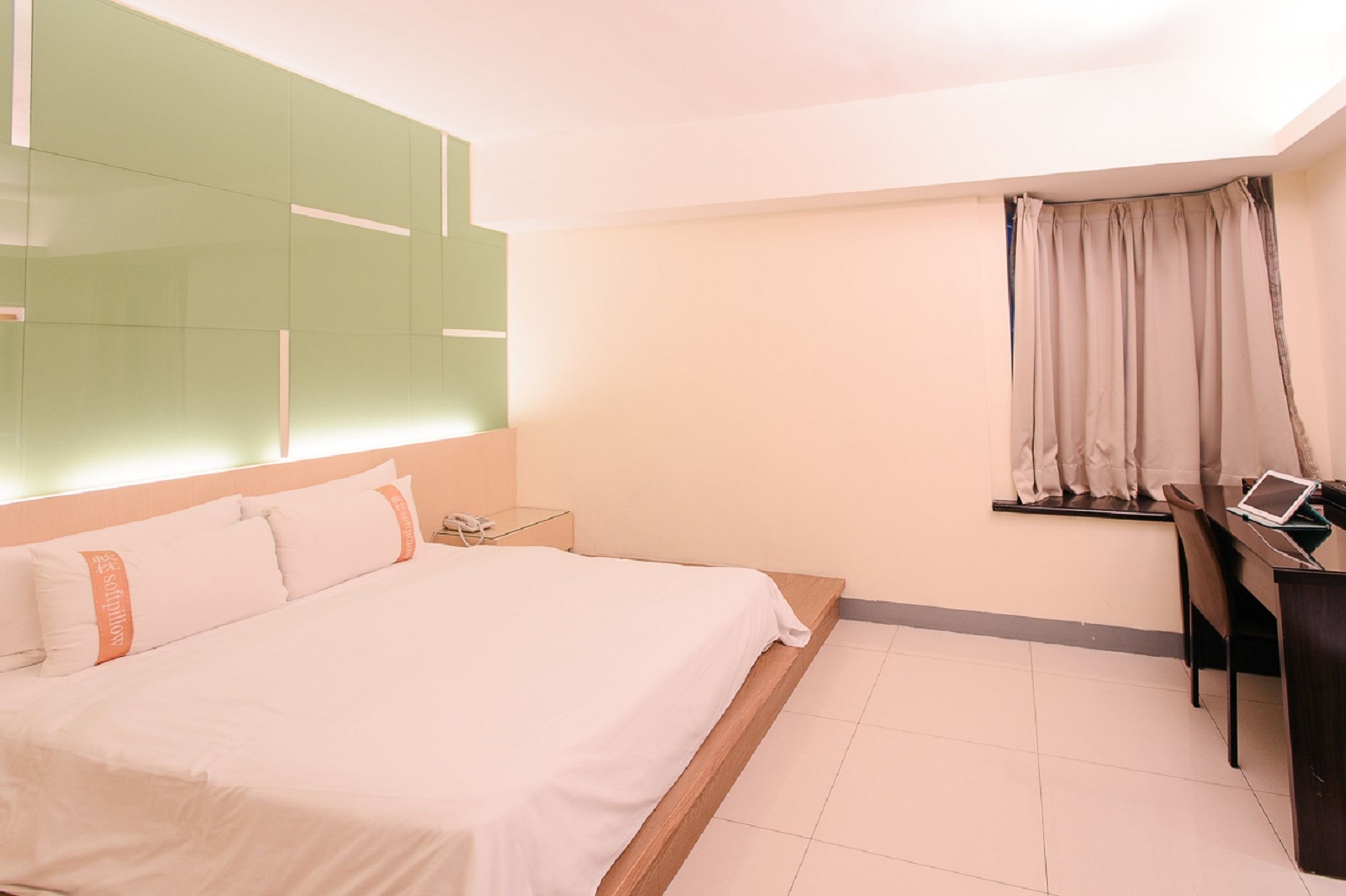 A hotel room with a large bed, a desk with a laptop, a bedside table with a phone, and a window with curtains.