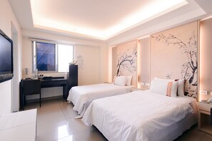 Habitación de hotel con dos camas individuales, un escritorio con silla, un televisor de pantalla plana, una ventana y un mural floral en la pared.