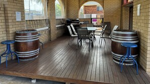 A covered patio with brick walls, a wooden deck, a table with chairs, and two wooden barrels. - Toreador Motel (Coffs Harbour)
