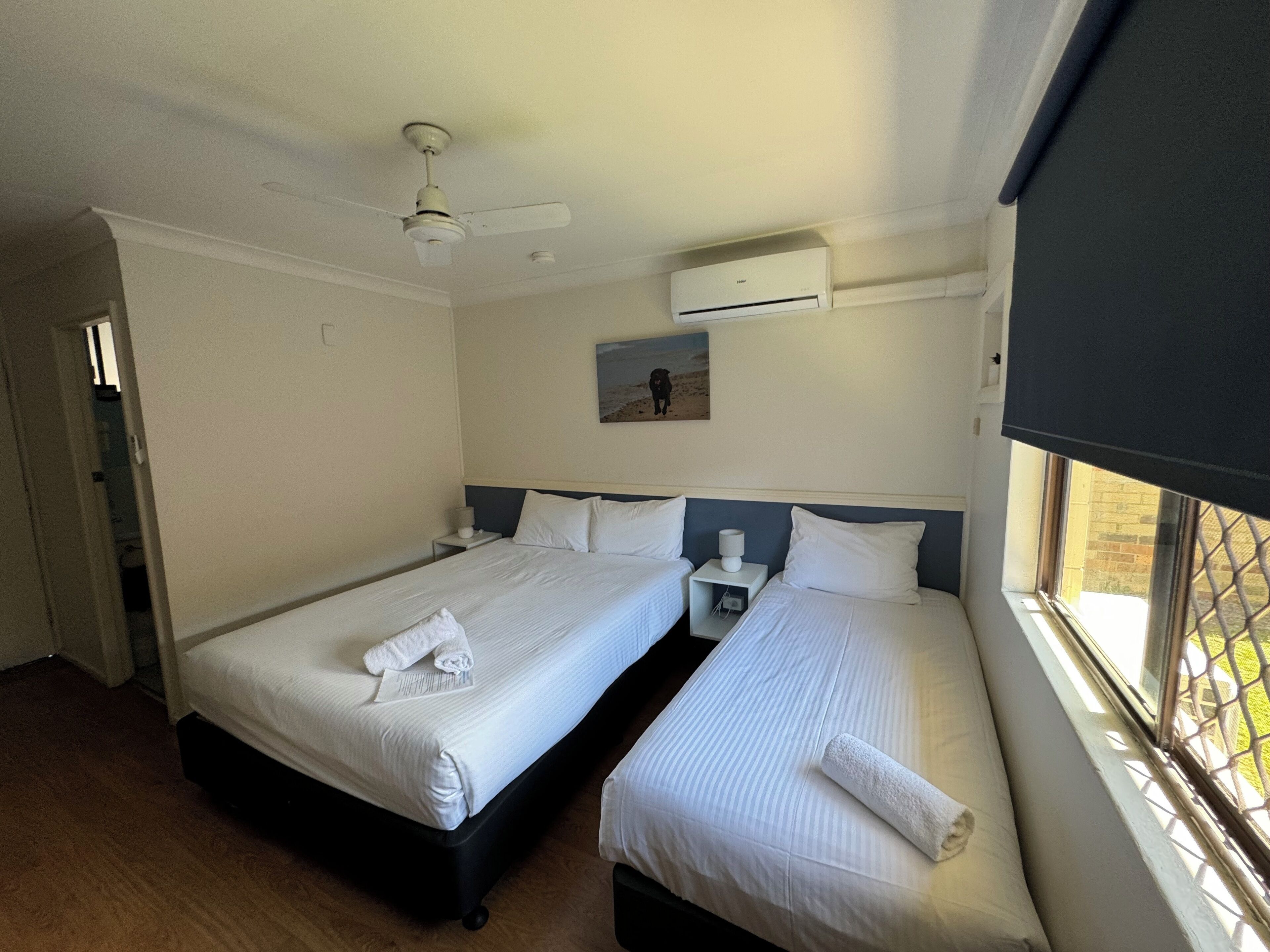A hotel room with two beds, a ceiling fan, an air conditioning unit, a painting on the wall, and a window with a view.