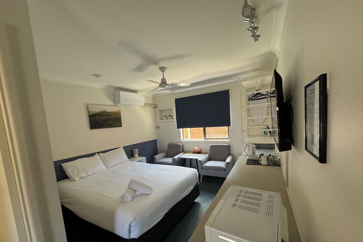 A hotel room with a bed, a desk, a chair, a ceiling fan, a window with blinds, and a TV mounted on the wall.