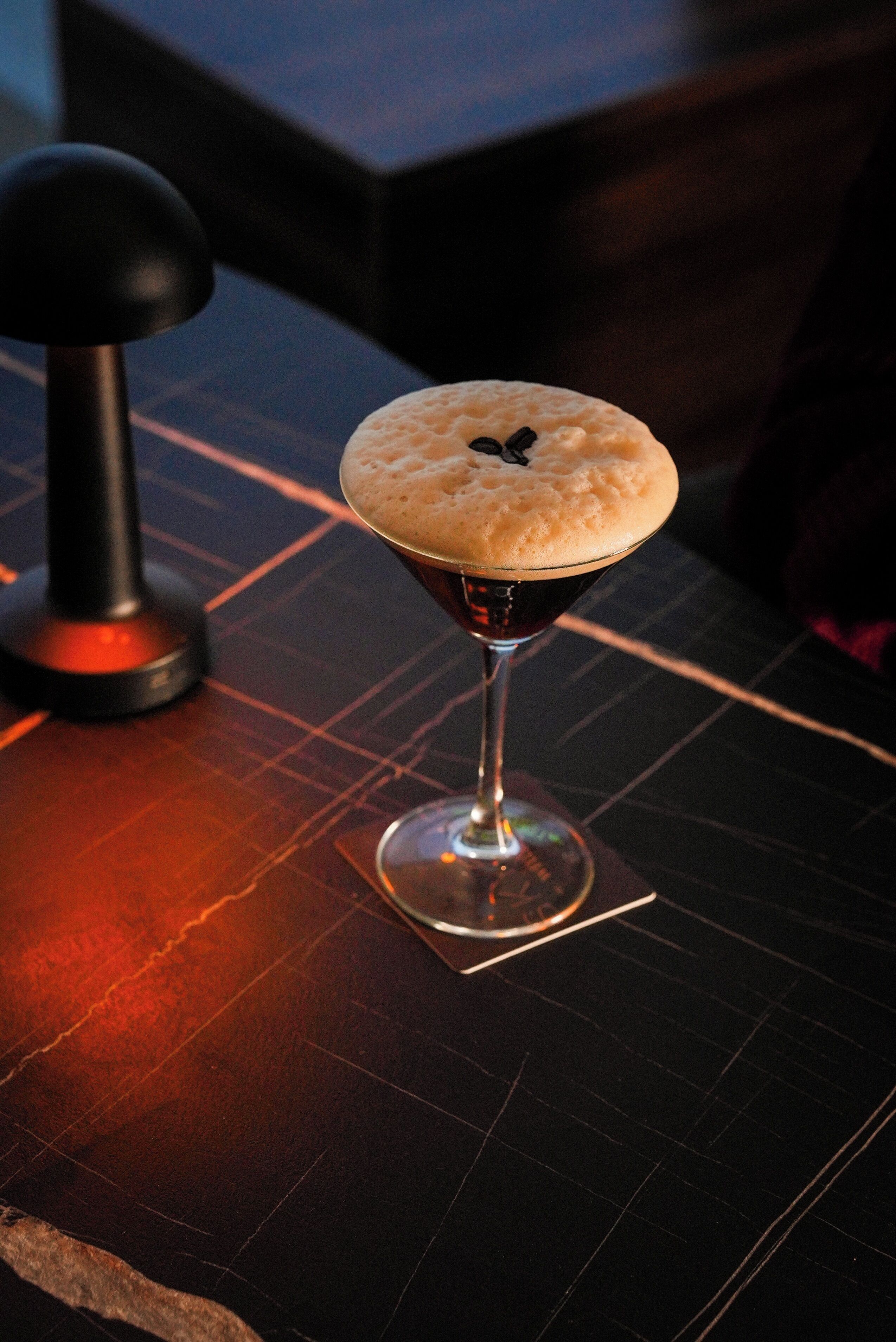 A cocktail in a martini glass with a frothy top, placed on a dark table with a geometric pattern.