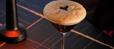 A cocktail in a martini glass with a frothy top, placed on a dark table with a geometric pattern.