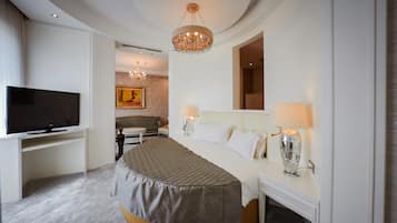 A bedroom with a large bed, a television, a chandelier, and a dining area in the background.