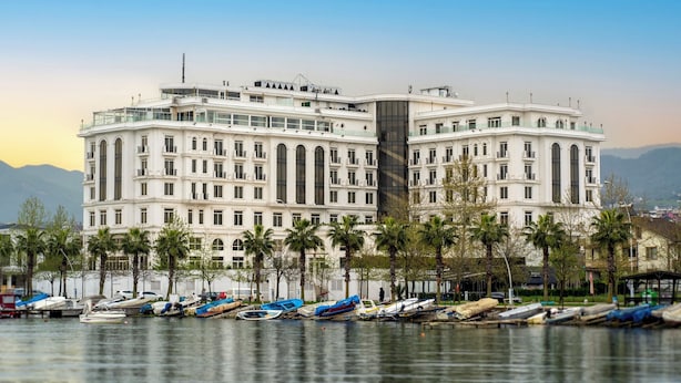 A large, multi-story hotel with a symmetrical facade, located by a body of water with several boats docked in front. - Wellborn Luxury Hotel (Basiskele)