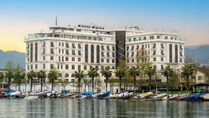 Un hotel grande de varios pisos con fachada simétrica, situado junto a un cuerpo de agua con varias embarcaciones amarradas.
