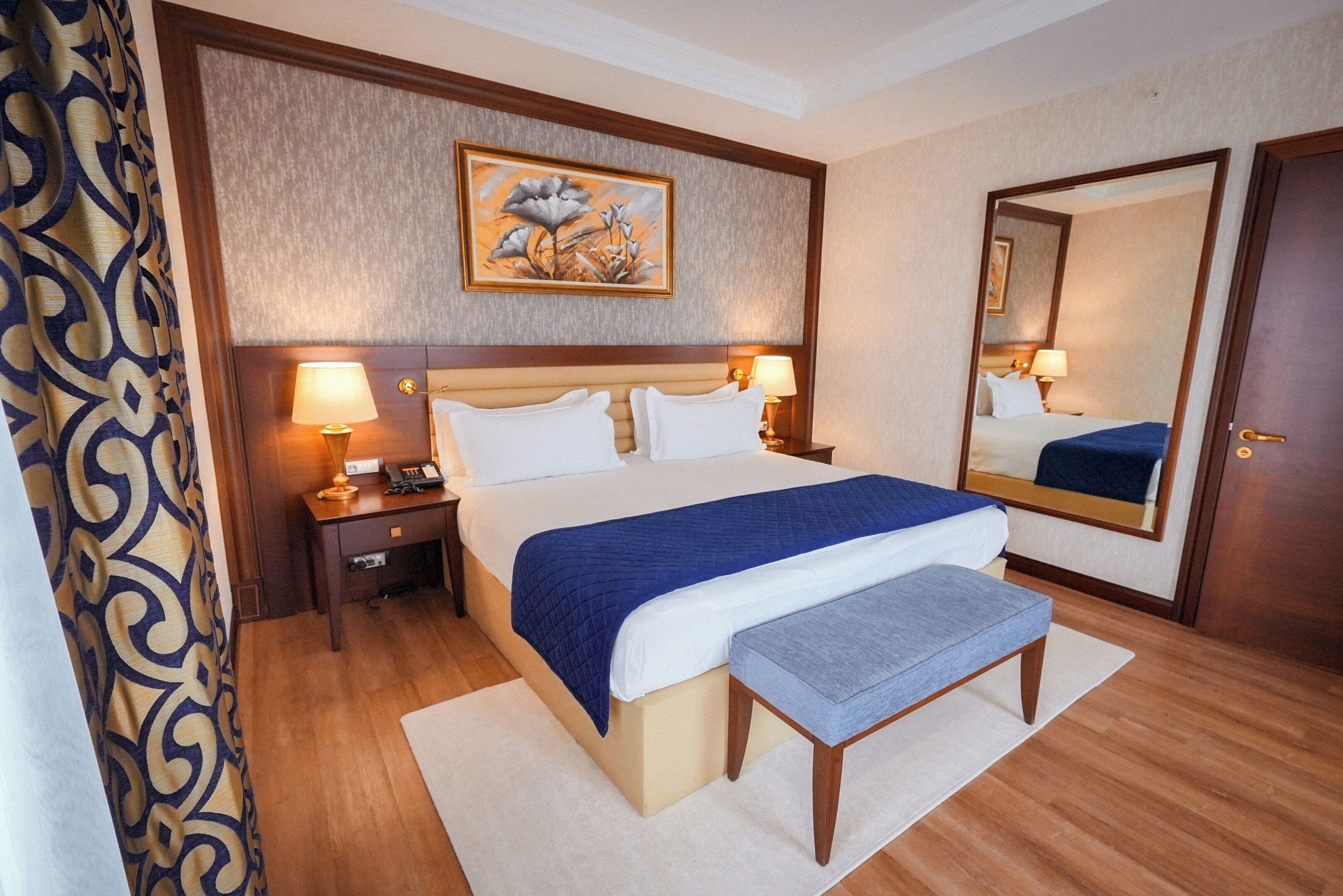 A hotel room with a bed, bedside tables, a nightstand, a bench, a large mirror, and a painting on the wall.