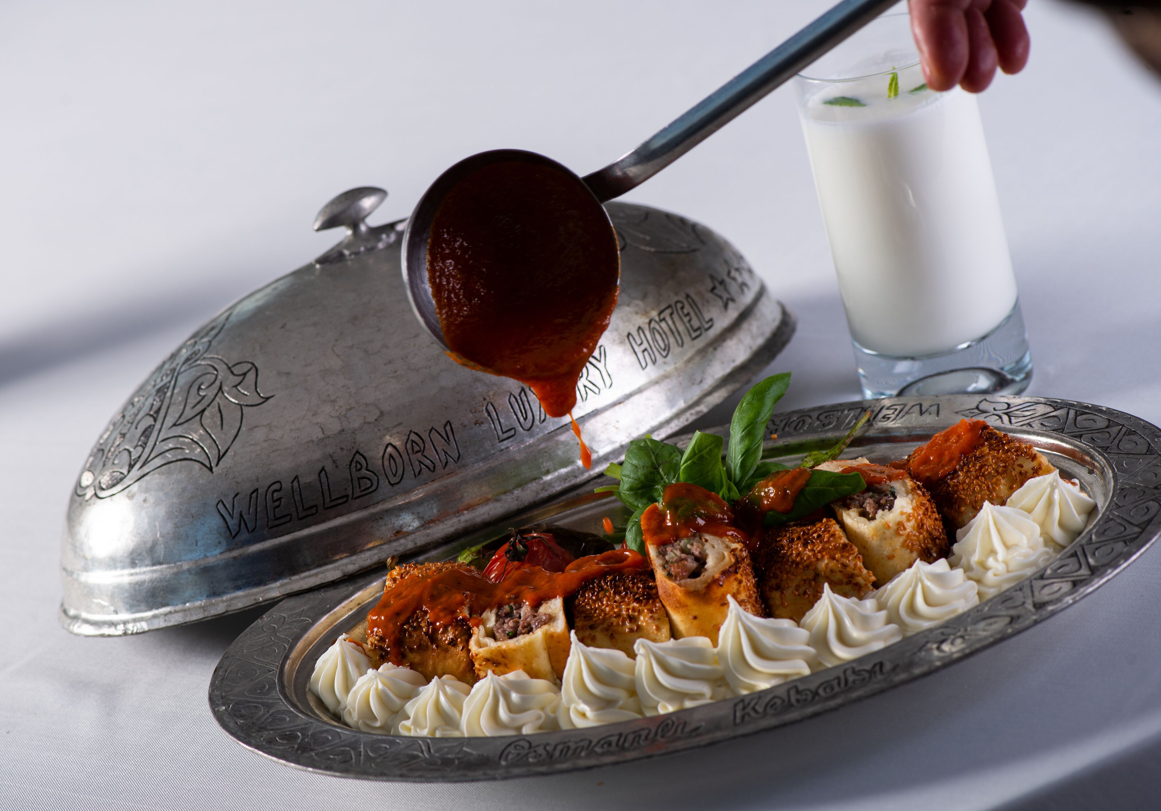A silver tray with a covered dish, a glass of milk, and a spoon pouring sauce over food.
