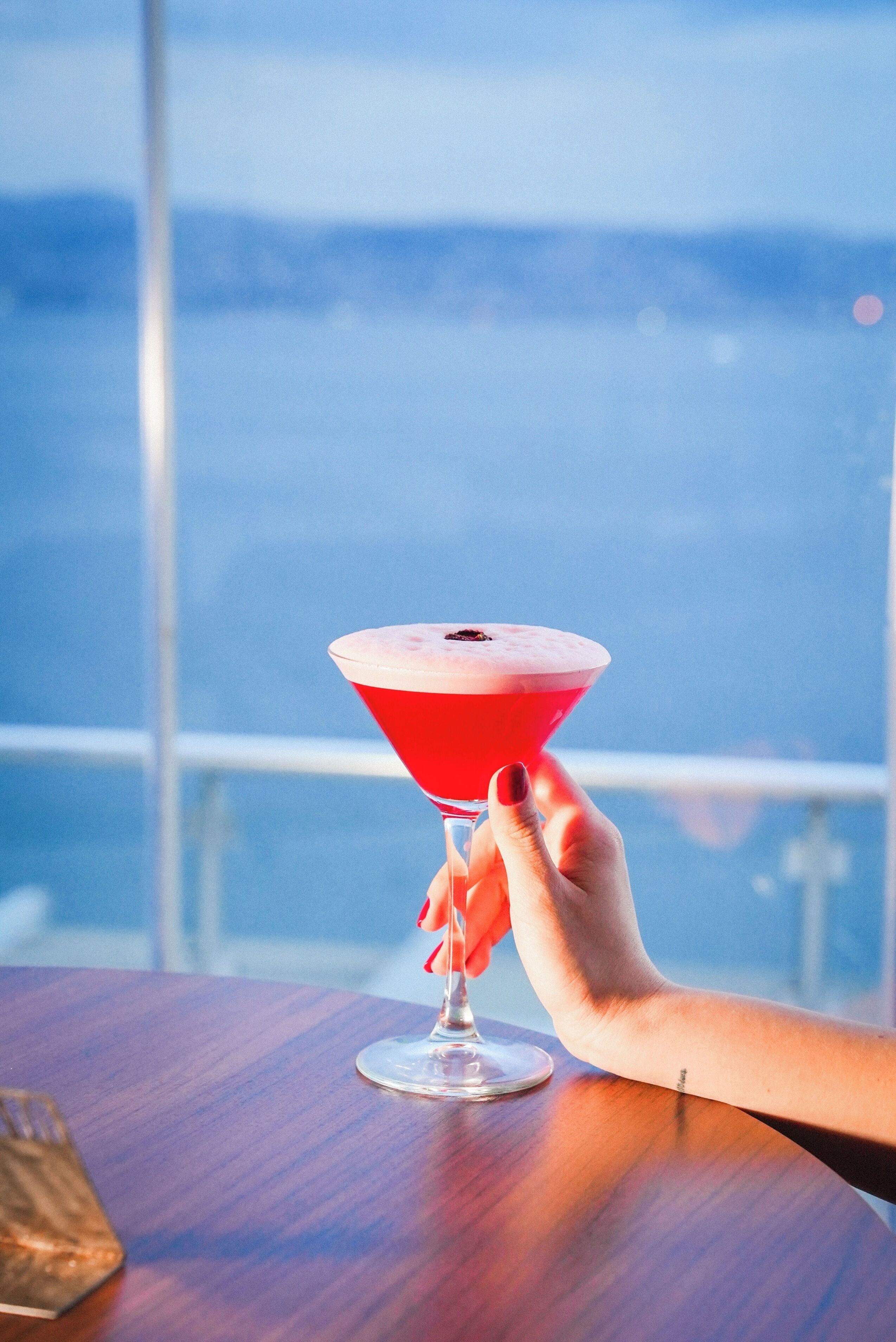 A hand holding a red cocktail glass on a wooden table with a view of a cityscape in the background.