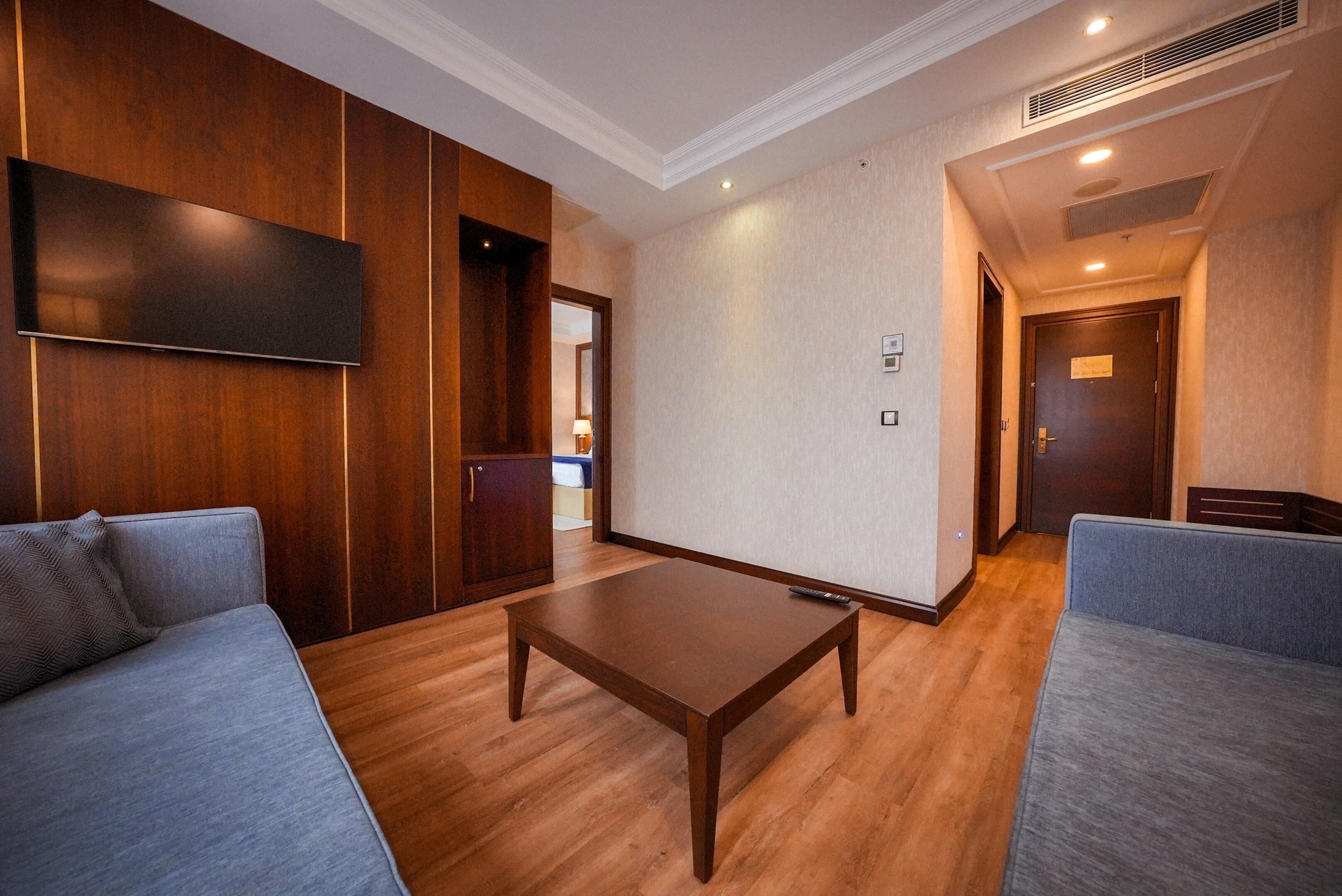 A hotel room with a wooden panel wall, a flat-screen TV, a wooden coffee table, and two grey sofas.