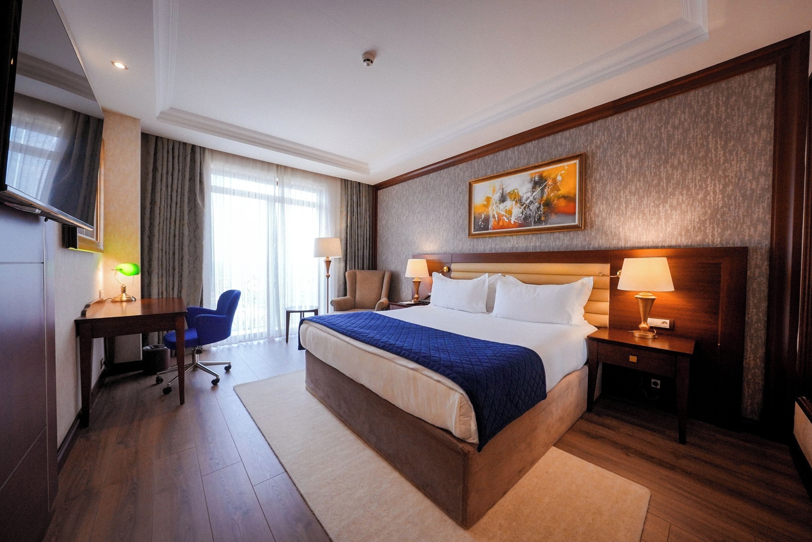 A hotel room with a large bed, a desk with a chair, a TV, and a painting on the wall.