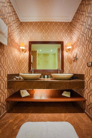 A bathroom with a large mirror, two sinks, a bench, and patterned wallpaper. - Wellborn Luxury Hotel (Basiskele)