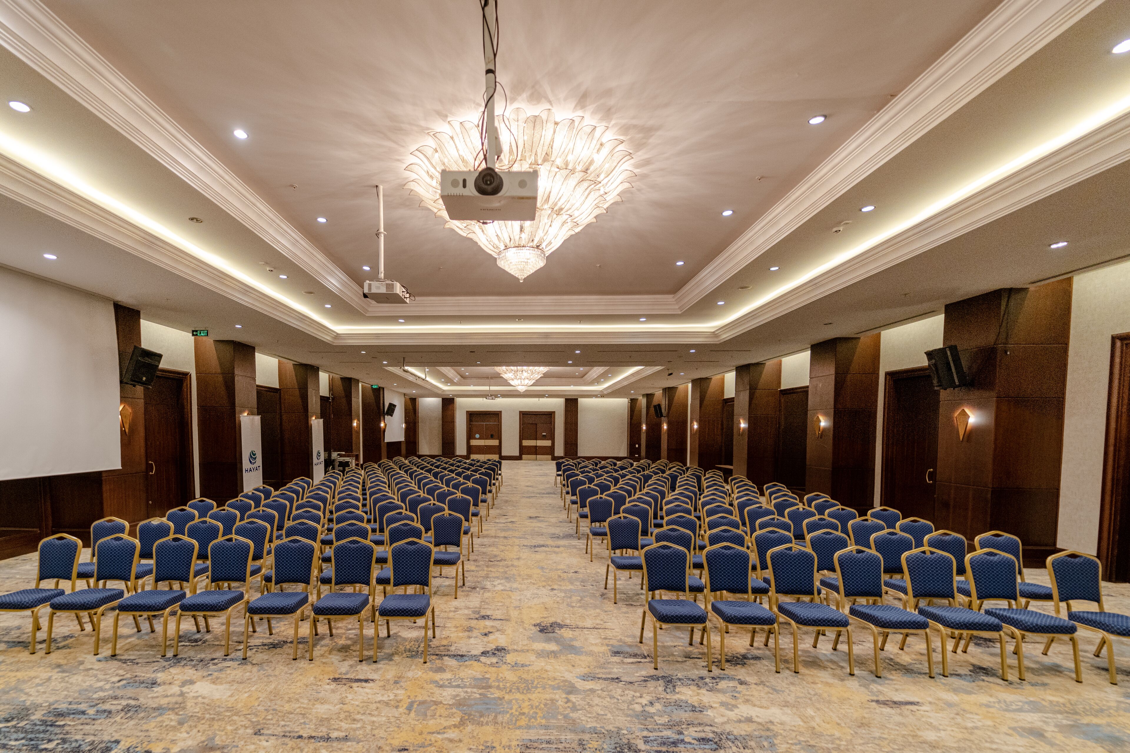 A conference room with rows of blue chairs, a chandelier, and a carpeted floor.