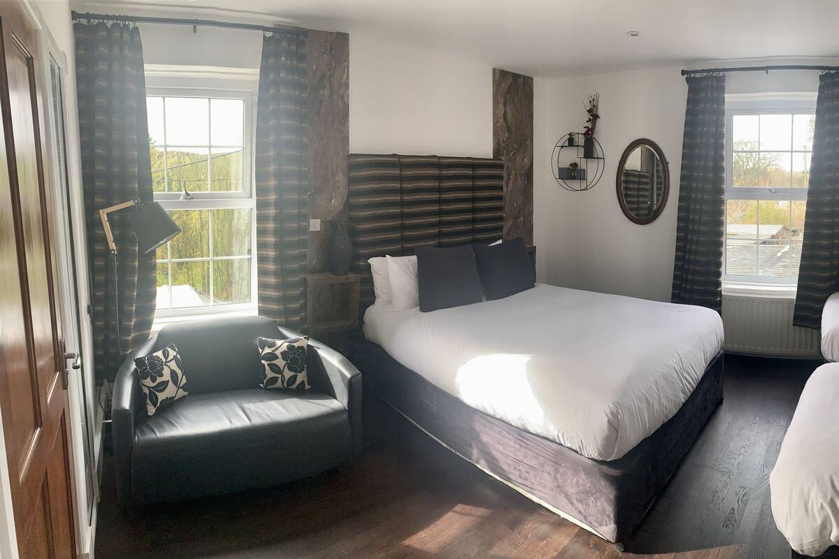A bedroom with a large bed, a smaller bed, a sofa, and a chair. There are two windows with curtains, a mirror on the wall, and a wooden headboard.