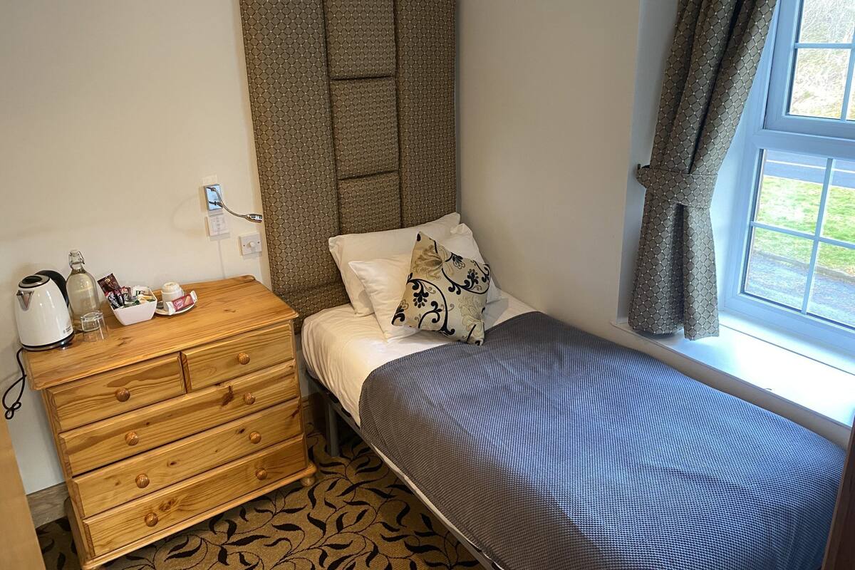 A small, neatly arranged bedroom with a single bed, a wooden dresser, a kettle, and a window with curtains.