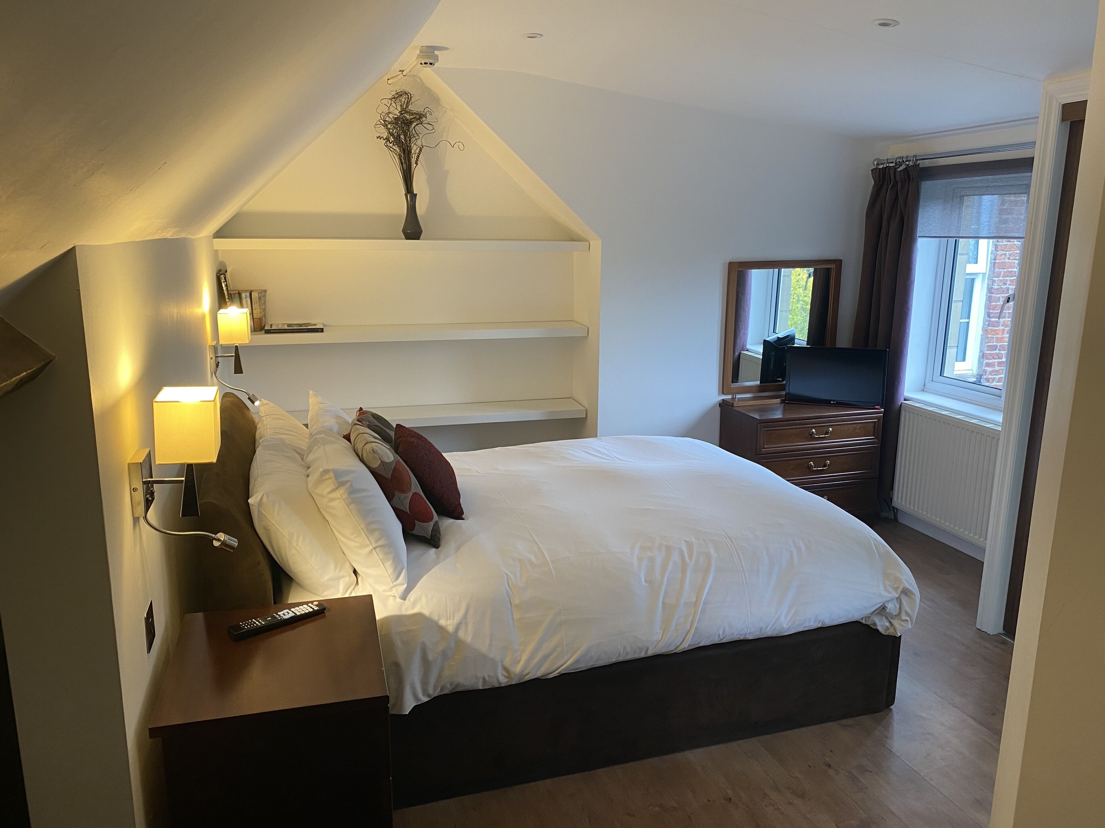 A hotel room with a bed, bedside lamps, a TV, a dresser, and a window with curtains.