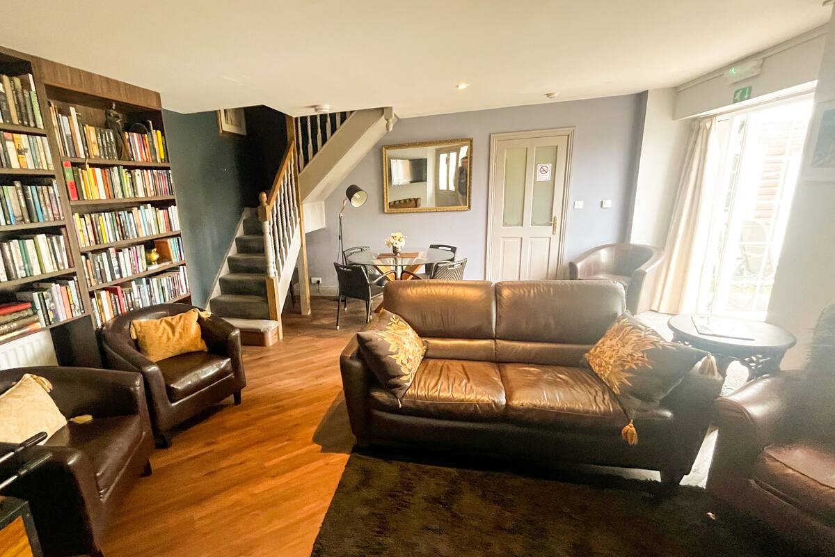 A living room with a leather sofa, armchairs, a bookshelf, a staircase, and a dining area.